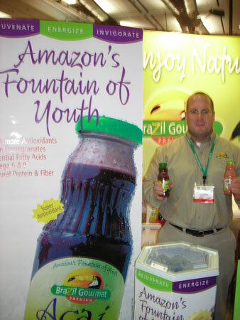 A man in a tan shirt and lanyard holds two small juice bottles beside a large Brazil Gourmet banner for an açaí drink labeled Amazon's Fountain of Youth.