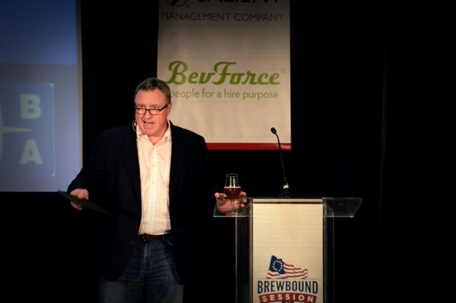 A man in glasses and a dark blazer stands at a clear podium holding a glass of beer, in front of a BevForce banner. The podium bears a Brewbound Session sign.