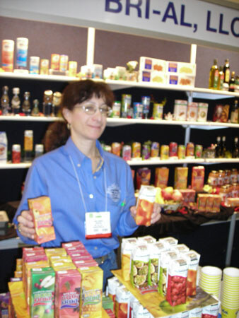 A woman with glasses and a blue shirt holds a boxed drink under a Bri-Al, LLC sign. Shelves and tables around her hold many colorful juice boxes and bottles.