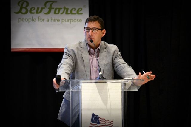 A man with glasses and a gray blazer stands at a clear podium holding a microphone in one hand and a pen in the other, with a BevForce banner behind him.