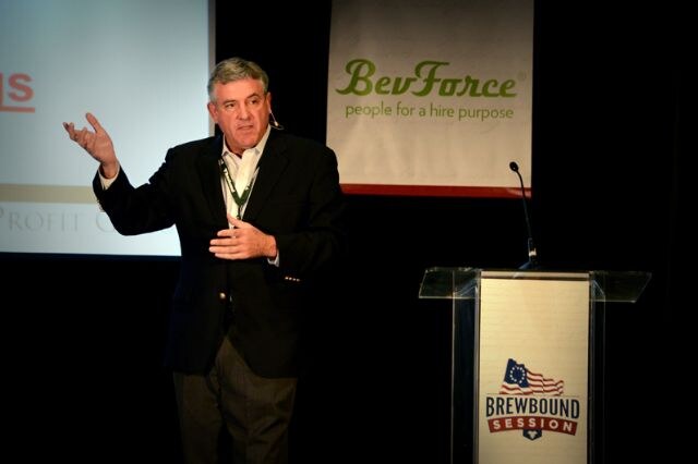 A gray-haired man in a dark blazer gestures while speaking on stage beside a clear podium marked Brewbound Session, with a BevForce banner and a projection screen behind him.
