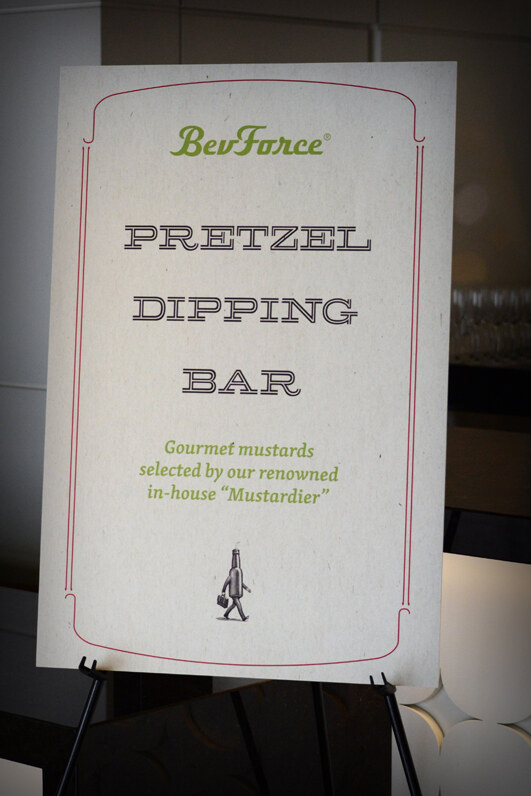 Easel sign reading BevForce Pretzel Dipping Bar, noting gourmet mustards selected by an in-house Mustardier, with a small illustration of a walking bottle carrying a briefcase.