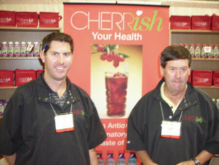 Two men in black Cherrish jackets with name badges stand in front of a red Cherrish banner showing a glass of cherry juice, with shelves of red boxes and bottles behind them.