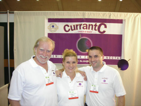 A bearded man, a woman and a man in white polo shirts stand with arms around each other in front of a purple Currant C banner.