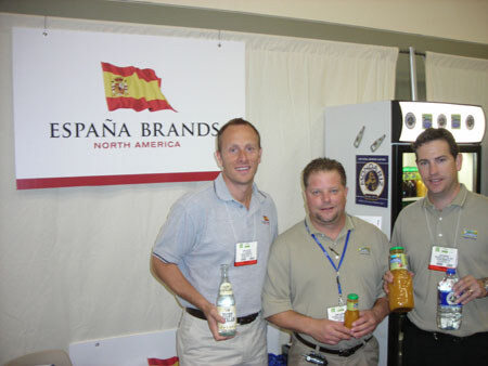 Three men in polo shirts and badges stand in front of an España Brands North America sign with a Spanish flag. Two hold bottled drinks and one holds a water bottle.