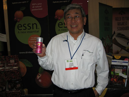 A smiling man with glasses and a white shirt holds up a small pink essn can in front of essn banners showing fruit graphics.