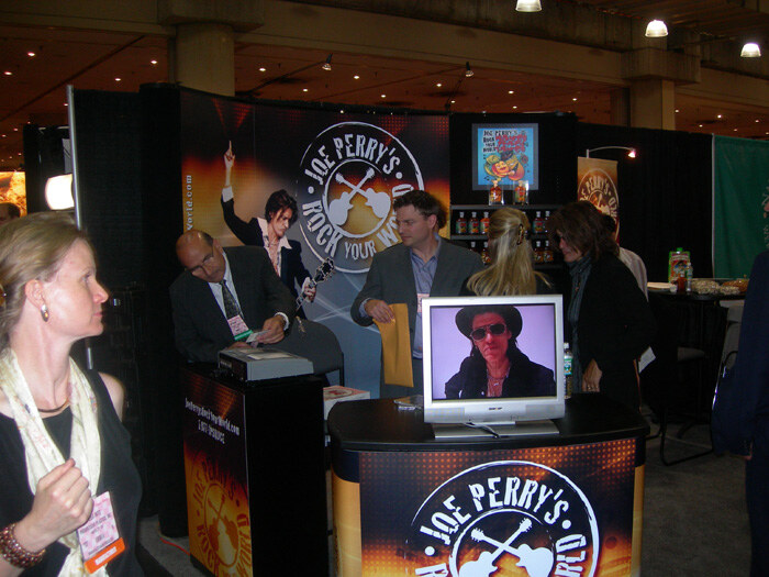 Visitors and staff gather in a dark booth with Joe Perry's Rock Your World signs. A monitor on the counter shows a person in a hat and sunglasses, and a man in a suit reads at a podium.