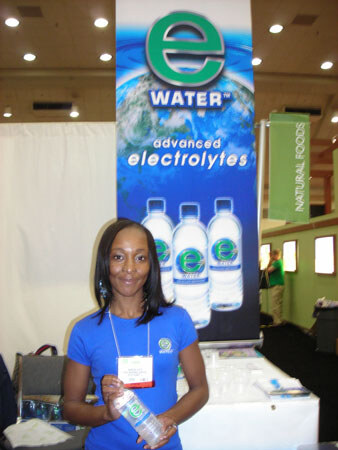 A woman in a blue shirt holds a bottle of e Water in front of a tall e Water banner reading advanced electrolytes, showing three bottles, at a trade show booth.