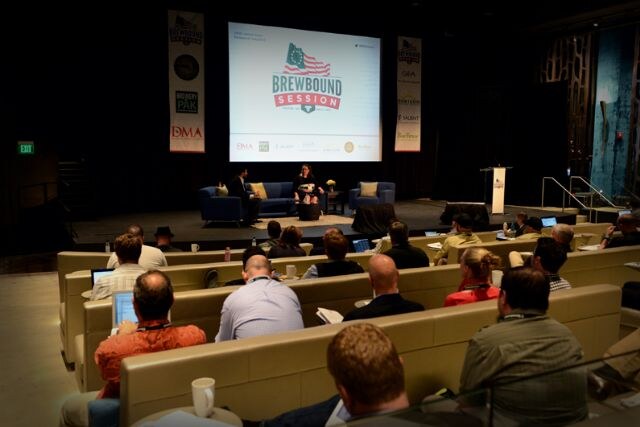 Seated attendees, some with laptops, face a stage where a few people sit on sofas below a screen reading Brewbound Session, with DMA and Brewery PAK banners hanging on either side.