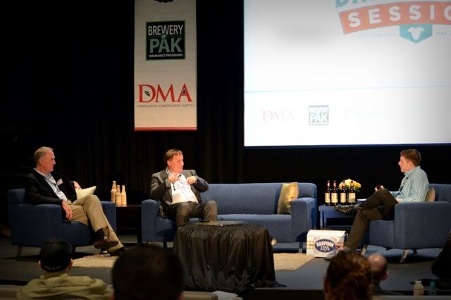 Three men sit on sofas and chairs on a stage for a panel, with bottles and a Harpoon IPA box on tables, Brewery PAK and DMA banners behind them and audience heads in the foreground.