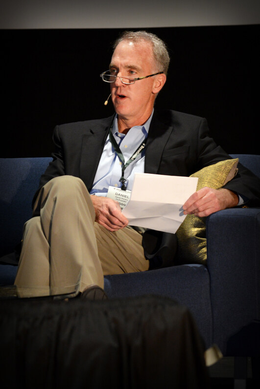 Daniel, a gray-haired man with glasses and a headset microphone, sits on a blue couch holding a sheet of paper and wearing a name badge.