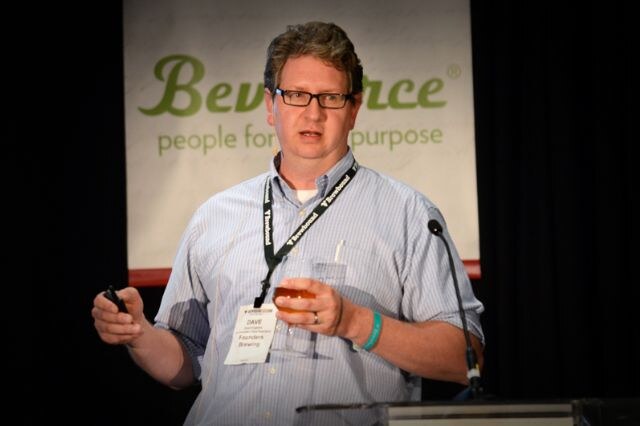 Dave of Founders Brewing, a man with glasses and a striped shirt, speaks near a microphone while holding a glass of beer and a clicker. A lanyard and name badge hang from his neck.