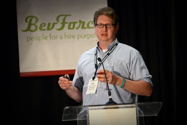 Dave of Founders Brewing stands at a clear podium holding a glass of beer and a clicker, in front of a BevForce banner. He wears glasses, a striped shirt and a lanyard with a name badge.