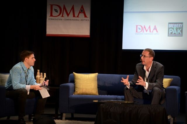 A man in a checked shirt holding papers and a microphone faces a man in a dark jacket who gestures while speaking, seated on a blue sofa beneath a DMA banner, with DMA and Brewery PAK logos on screen.