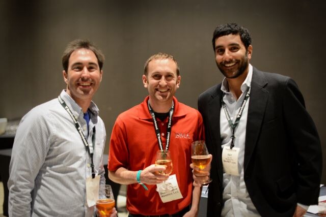 Three smiling men with lanyards hold glasses of drink; the middle man wears a red polo with a DMA logo.