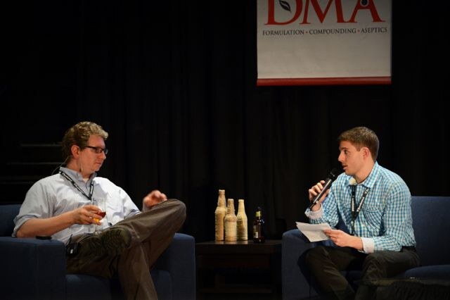 Two men sit in armchairs on a dark stage under a DMA banner. The man on the left holds a glass of beer; the man on the right wears a blue plaid shirt and speaks into a microphone while holding papers.