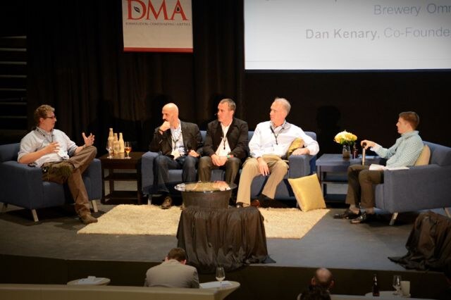 Five men sit in armchairs and on a sofa on a stage for a panel discussion, with a DMA banner behind them. The man on the left gestures while speaking. A bottle display sits on a side table.