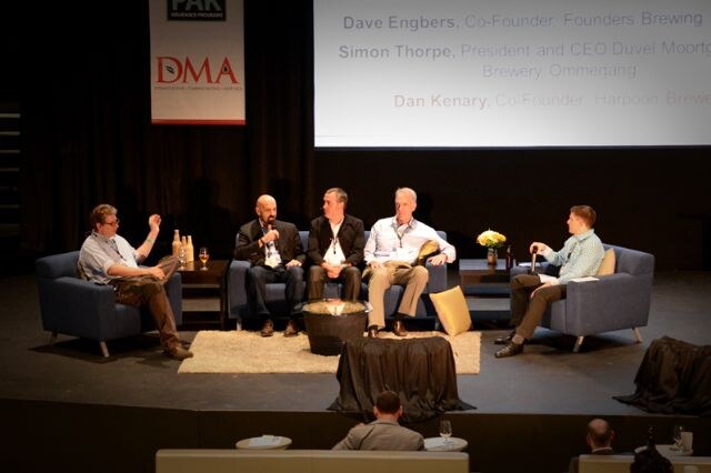 Five men sit on stage in a panel setting beneath a DMA banner, with a projected slide of names and titles above them. The man at far left gestures with one hand; another holds a microphone.