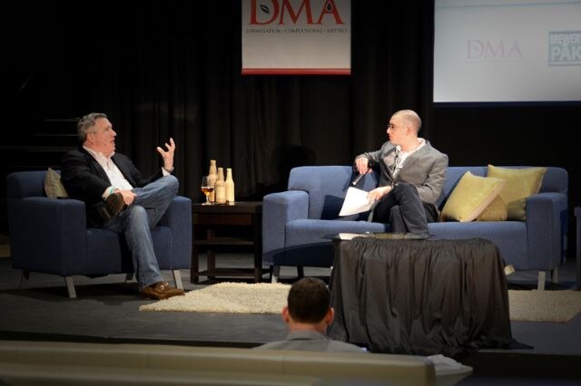A man in a dark jacket and jeans gestures while seated in an armchair, facing a man in a gray jacket holding papers on a sofa. A DMA banner hangs behind them, and an audience member is in the foreground.