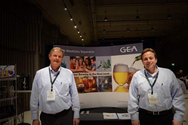 Two men in light blue button-up shirts and lanyards stand in front of a GEA Westfalia Separator display showing beer glasses; Fred is the man on the right.