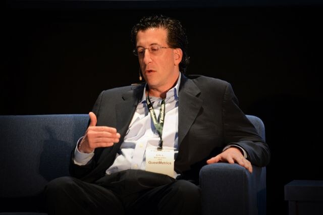A man with glasses and a headset microphone, wearing a dark blazer and a GuestMetrics badge, gestures while seated in a blue armchair on a dark stage.