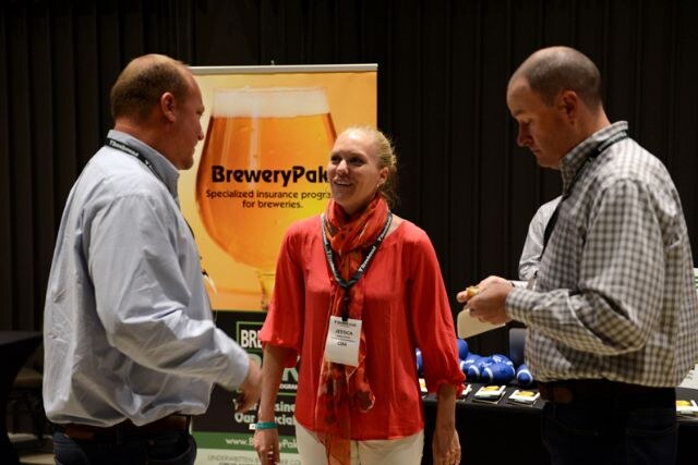 Jessica, in an orange top and scarf, smiles at the center between two men in conversation in front of a BreweryPak banner showing a glass of beer.