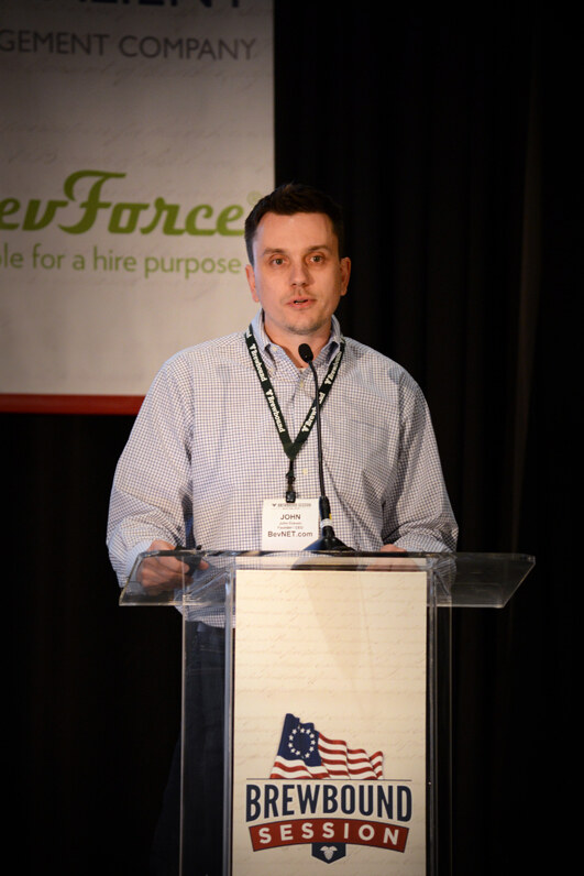 John of BevNET.com stands at a clear Brewbound Session podium speaking into a microphone, wearing a checked shirt and lanyard, with a BevForce banner behind him.