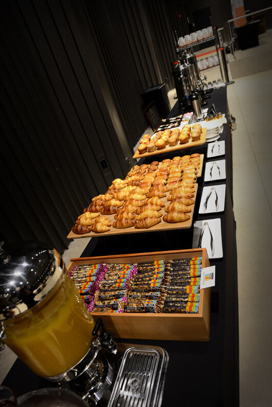 Buffet table with trays of croissants and muffins, a wooden box of KIND bars, serving tongs and a glass dispenser of orange juice in the foreground.