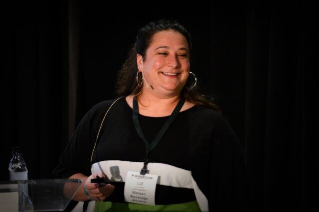 Liz, a smiling woman with dark hair, hoop earrings and a badge on a lanyard, holds a clicker and wears a headset microphone while speaking against a dark curtain.