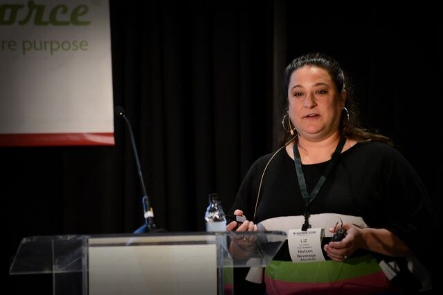 Liz, wearing a badge lanyard and a black top with colored stripes, speaks at a clear podium with a microphone, holding a small clicker, in front of a black curtain.