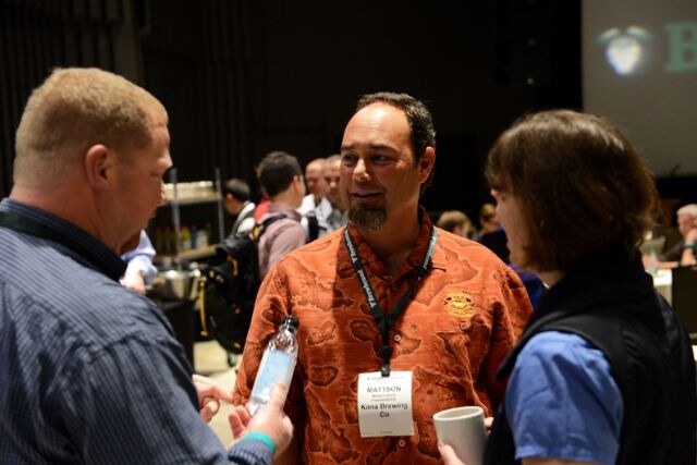 Mattson of Kona Brewing Co, wearing an orange patterned shirt and badge, talks with two attendees in the foreground while others mingle behind.