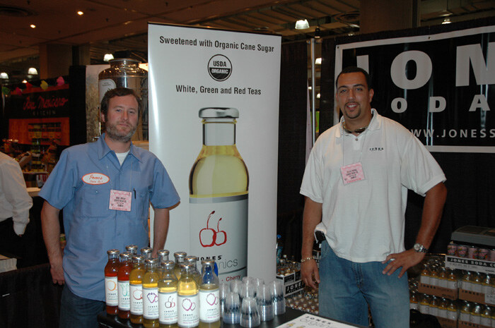 Two men stand beside a table of Jones Soda bottles, one in a blue work shirt and one in a white polo, in front of a banner with organic cane sugar tea bottles and a Jones Soda sign.