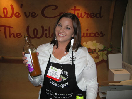 A smiling woman with dark hair wears a white shirt and a black Fruit Technologies apron with a name badge. She holds up a bottle of amber juice in front of a wall with large script lettering.