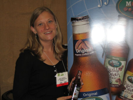 A smiling woman with blonde hair holds a small bottle in front of a large Green Mountain Original organic coffee soda graphic and Double Bean Elixir bottles.