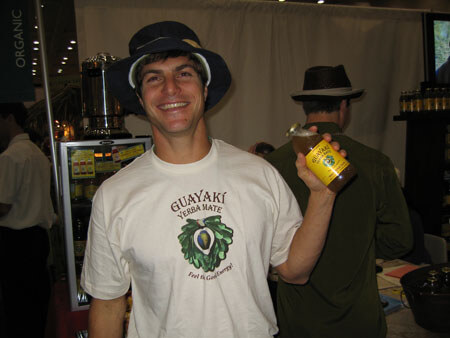 A smiling man in a dark hat and a white Guayaki Yerba Mate t-shirt holds up a yellow bottle. Another person in a hat stands behind him in the exhibit hall.