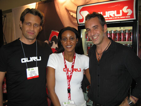 Two men and a woman pose together at a Guru booth. The woman wears a Guru lanyard and shirt, and a Guru-branded cooler of energy drinks is behind them.