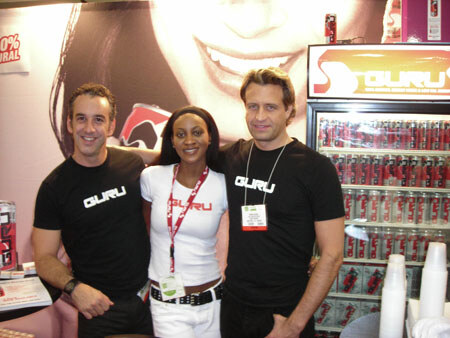 Two men and a woman in black and white Guru shirts pose together in a booth. A refrigerated cooler of red Guru cans stands behind them at right.