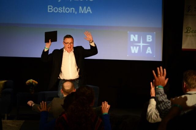 A man in a dark blazer and glasses raises both arms, holding a tablet in one hand, in front of a screen showing the NBWA logo and Boston, MA. Audience members in the foreground also raise their hands.