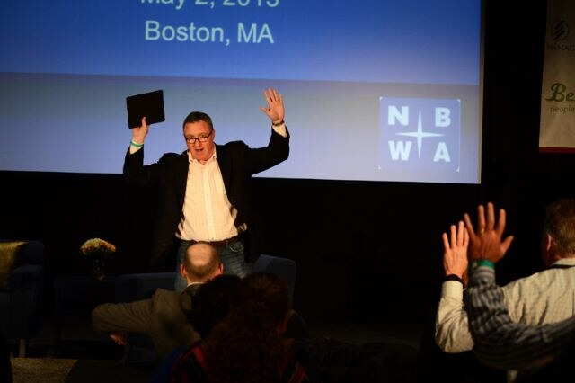 A man in a dark blazer and glasses holds a tablet and raises both hands before a screen with the NBWA logo and Boston, MA. Several audience members in the foreground raise their hands.