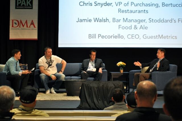 Four men sit on stage in a panel discussion, one gesturing. A screen lists speakers and a DMA and PAK Insurance Programs banner hangs at left, with audience heads in front.