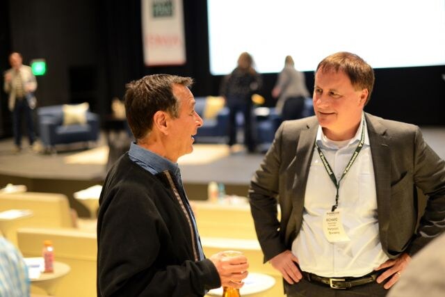 Richard, wearing a gray blazer and a badge on a lanyard, stands at right talking with a man in a dark jacket holding a bottle, in a seating area with a stage behind them.