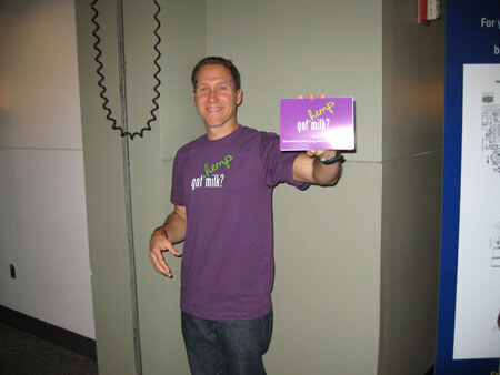 A smiling man in a purple hemp got milk? t-shirt holds up a small purple box of the same brand in an indoor hallway.