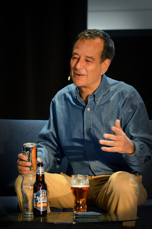 A man in a denim shirt sits on a couch holding a Boston Lager can, with a Samuel Adams bottle and a glass of beer on the table in front of him.