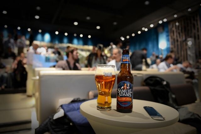 A bottle of Samuel Adams Boston Lager and a filled glass sit on a small round table with a phone, with a blurred seated audience in the background.
