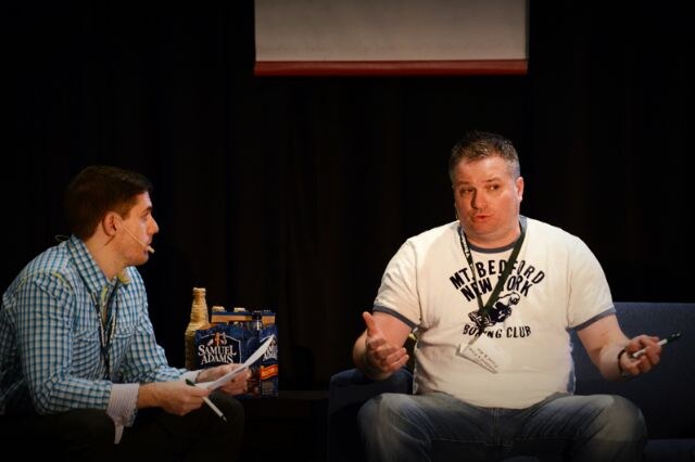 On stage, a man in a blue checked shirt holding papers faces a man in a Mt. Bedford New York Boxing Club T-shirt; a Samuel Adams six-pack sits on the table between them.