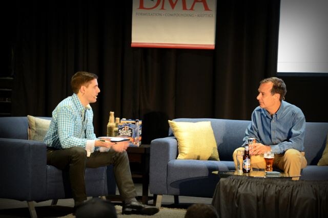 Two men sit on blue couches on stage, one with papers and one holding a beer, with a Samuel Adams six-pack between them and a DMA banner behind.