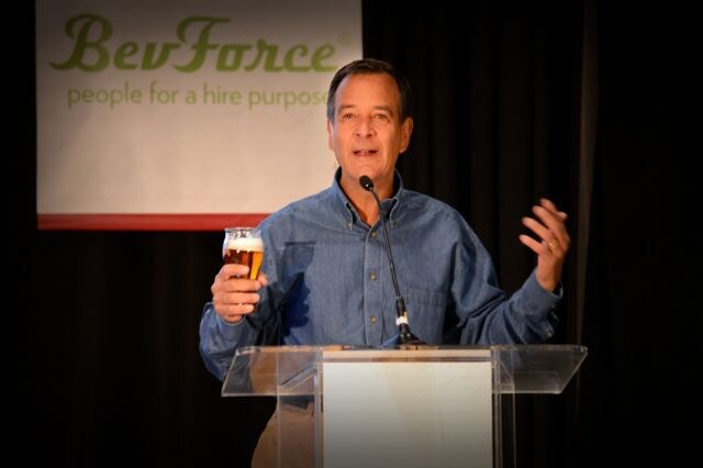 A man in a denim shirt holds a glass of beer and gestures while speaking at a clear podium with a microphone, in front of a BevForce banner.