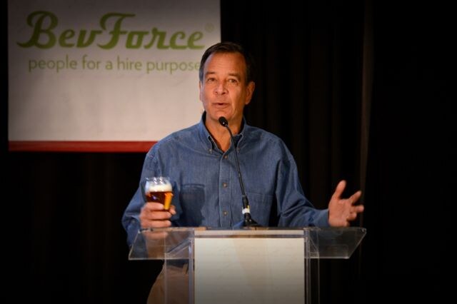 A man in a denim shirt holding a glass of beer speaks at a clear podium with a microphone, with a BevForce banner behind him.