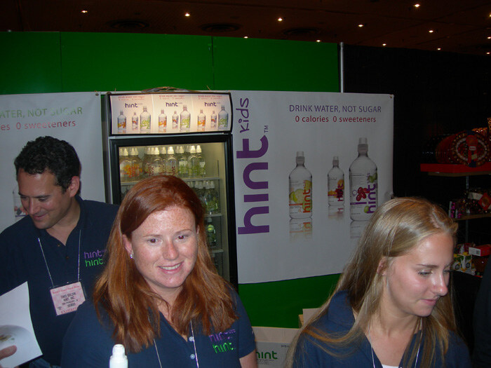Three people in navy Hint shirts stand at a booth with a Hint Kids banner reading 'Drink water, not sugar' and a lit refrigerated display case of bottled water behind them.