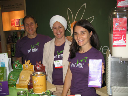 A man, a woman in a white turban and a woman in purple hemp got milk? shirts stand at a booth with product tins, boxes and a hempmilk dispenser; one woman holds a purple carton.